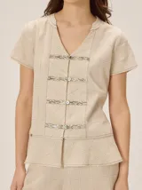 Camisa de manga corta color beige con estampado a cuadros, cuello en V y detalles de galones decorativos estilo oriental en la parte frontal.