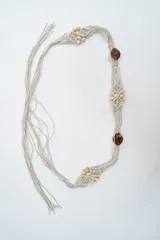 Cinturón de macramé color beige, con diseño trenzado artesanal, decorado con cuentas de madera claras y piezas circulares de resina color ámbar.