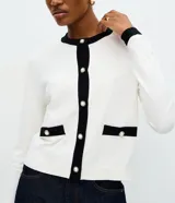 Cardigan de punto color blanco con detalles en negro en cuello, puños y tapeta frontal. Presenta cierre frontal con botones perlados y dos bolsillos delanteros falsos con solapa bicolor.