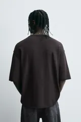 Camiseta negra de algodón con corte boxy fit y cuello redondo. Presenta un estampado gráfico desgastado con temática espacial o celestial en tonos azules y blancos, y texto en la parte superior e inferior.