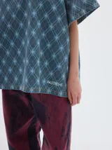 T-shirt oversize de denim con estampado de rombos en tonos azules, cuello redondo y mangas cortas. Presenta un bordado de estrellas blancas en el pecho y costuras visibles en color marrón claro.
