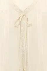 Camisa de gasa color crudo con escote en pico, manga corta abullonada con terminación en volante y cierre frontal con botones ocultos y lazada. Presenta delicados detalles de bordados a tono en toda la prenda.
