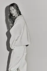 Campera de tejido suave efecto peluche en color crudo, con cuello redondo, hombros caídos y cierre frontal mediante alamares con detalles en contraste negro. Cuenta con bolsillos laterales ribeteados y forro interior.