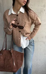 Cardigan de punto trenzado en color beige, con cuello redondo, cierre frontal de botones oscuros y mangas largas con puños acanalados.