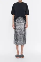 Falda midi plateada con lentejuelas y flecos en el ruedo, corte recto, cierre trasero con cremallera invisible y abertura frontal.