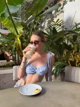 Top corto de estilo bralette con estampado de rayas verticales finas en color celeste y blanco. Presenta un diseño fruncido en el centro del busto y breteles finos que se atan en los hombros.