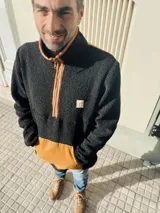 Buzo de hombre de polar negro con cierre parcial color naranja, bolsillo de parche color naranja en la parte inferior y cuello alto.