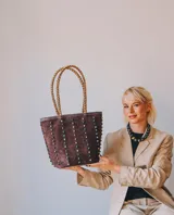 Cartera tipo tote de gamuza color marrón, con correas trenzadas y detalle de piedras naturales incrustadas en líneas verticales. Cuenta con cierre metálico y forro interior de tela.