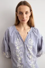 Blusa de manga larga con estampado de rayas verticales en tonos celestes y blancos. Presenta un escote en V con cierre frontal de botones y detalles de bordados florales en color crema en el frente y en los puños.
