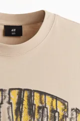 Remera de algodón en color beige con cuello redondo acanalado, hombros caídos y corte holgado. Presenta un estampado frontal con la frase 'RUN WILD' en tonos amarillos y negros con efecto desgastado.