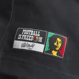 Remera de algodón negra con cuello redondo y mangas cortas. Presenta el logo del trébol de Adidas bordado en blanco en el lado derecho del pecho y un parche circular con los colores rasta en el lado izquierdo. Incluye un parche rectangular en la parte inferior con la inscripción Football is Freedom y una imagen de Bob Marley.