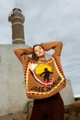 Sweater tejido de lana color marrón con cuello redondo y puños ajustados. Presenta un diseño con un sol amarillo, un surfista negro y la inscripción "José Ignacio" en el frente.