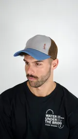 Gorra tipo trucker con frente gris, visera azul y panel trasero de malla marrón. Presenta un pequeño bordado en el frente.