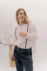 Camisa de algodón blanca con cuello a la base, cierre frontal con botones y mangas largas con puños delicados. Presenta volados verticales y detalles de cintas de terciopelo en contraste a lo largo del frente.