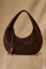 Cartera tipo hobo de gamuza color marrón oscuro, con diseño curvado, asa de hombro integrada y cierre con tirador de cuero decorativo.