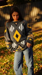 Sweater tejido a mano con técnica de intarsia y detalles bordados, de diseño holgado y cuello redondo. Presenta un patrón geométrico de rombos en tonos gris, amarillo, blanco y marrón.