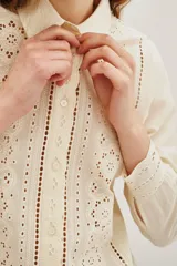 Camisa de algodón color crudo con diseño de encaje calado y detalles de plisados verticales en el frente. Presenta cuello clásico, cierre frontal de botones y mangas largas con puños delicados.