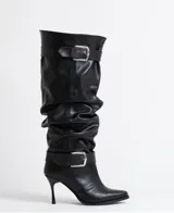 Bota alta de caña slouchy en color negro, confeccionada en cuero sintético. Presenta un diseño con efecto arrugado, puntera fina y tacón de aguja. Incluye dos correas ajustables con hebillas metálicas plateadas, una ubicada en la parte superior de la caña y otra en la zona del tobillo.