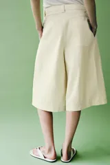 Bermudas de tiro medio color amarillo claro, confeccionadas en mezcla de algodón. Presentan cintura con trabillas, bolsillos laterales ocultos en costura, pliegues delanteros y cierre frontal con cremallera, botón y ganchos metálicos.