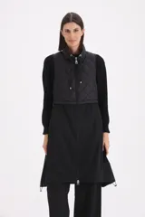 Chaleco largo negro de diseño combinado, con parte superior en matelassé y parte inferior de tela lisa. Presenta cuello alto con cordones ajustables y cierre frontal completo.