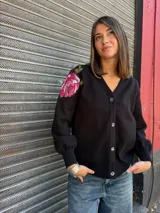 Cárdigan negro de algodón con escote en V y botones frontales. Presenta un aplique bordado de una rosa fucsia con hojas verdes en el hombro izquierdo.