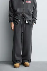 Pantalón jogger de corte wide fit confeccionado en tejido de algodón con interior de rizo. Presenta cintura elástica ajustable con cordón, bandas laterales decorativas, bolsillos laterales y un bolsillo trasero de plastrón. El acabado presenta un efecto lavado que le otorga una apariencia única.