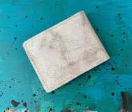 Billetera compacta blanca con diseño artístico pintado a mano en tonos turquesa, marrón y negro, perteneciente a la línea Ottenta.