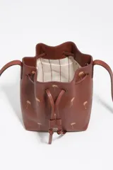 Bolso tipo bucket Bimba y Lola modelo Carlino, confeccionado en cuero marrón con estampado de pequeñas palmeras doradas y logo impreso en la parte frontal. Cuenta con cierre de cordón ajustable y asas largas para llevar al hombro.