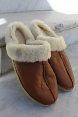 Pantufla de cuero color habano con interior de piel sintética, suela de goma y detalle de costura central.
