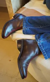 Botines de cuero color marrón oscuro con costuras rojas a mano en el borde superior. Tienen taco bajo y punta media.