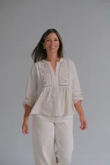 Blusa de color beige con cuello mao y cierre frontal de botones. Presenta detalles de bordado floral y calados en el frente, con mangas largas de corte holgado.