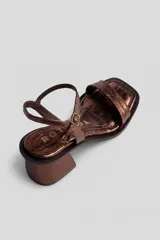 Sandalia de taco medio y ancho, confeccionada en cuero con acabado metalizado. Presenta una tira delgada en la puntera y tira ajustable con hebilla alrededor del tobillo.