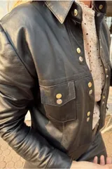 Camisa corta de cuero color marrón con cuello clásico, cierre frontal con botones metálicos dorados y plateados, bolsillos de parche en el pecho y corte a la cintura.