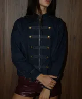 Chaqueta de denim azul oscuro con estilo militar, cuello alto, cierre frontal con cremallera y detalles de trabas decorativas con botones dorados.