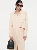 Camisa cropped de fit clásico color beige, con cuello camisero, botones frontales y un bolsillo en el pecho. Tiene mangas largas con puños abotonados y un largo trasero asimétrico.