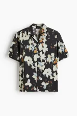 Camisa H&M de manga corta y corte regular fit, con cuello abierto y cierre frontal de botones. Presenta un diseño estampado de flores abstractas en tonos blancos, celestes y ocres sobre base negra.