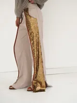 Pantalón acampanado de tiro medio, confeccionado en tela brocada con estampado floral en tonos beige y dorado, con franjas laterales de lentejuelas doradas y ribetes de cuero marrón.