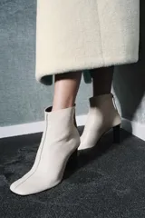 Botín de cuero color crema con puntera cuadrada y cierre de cremallera en la parte trasera. El tacón es ancho y de color negro. La altura del tacón es de 8,5 cm.