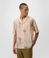 Camisa de lyocell color beige con corte boxy, cuello bowling y mangas cortas. Presenta un delicado bordado floral artesanal y cierre frontal con botones.