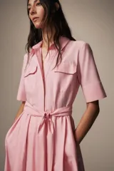 Vestido midi de estilo camisero color rosado, con cuello solapa, manga corta y cierre frontal con botones. Presenta bolsillos de plastrón con solapa en el pecho, bolsillos laterales ocultos y un cinturón del mismo tejido para ajustar en la cintura.