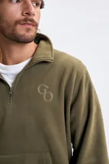Buzo de polar verde militar con medio cierre frontal, cuello alto y bolsillo tipo canguro. Presenta un pequeño logo bordado en el pecho.