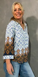 Blusa de manga larga con cuello camisero, corte recto y cierre frontal de botones. Presenta un diseño estampado geométrico en tonos celeste, blanco y marrón.
