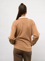 Buzo de felpa color tostado con cuello alto y medio cierre frontal. Presenta un diseño de corte holgado con hombros caídos, puños y ruedo acanalados, y el logo de la marca bordado en el pecho.