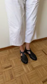 Sandalia de cuero marrón con diseño trenzado artesanal en el empeine, pulsera ajustable al tobillo con hebilla metálica y suela plana.