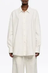 Camisa de corte recto en color blanco, con cuello clásico y cierre frontal de botones. Presenta mangas largas con puños abotonados y una textura sutil en el tejido.