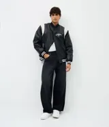 Campera bomber de cuero sintético con diseño bicolor, cuerpo negro y mangas blancas. Presenta cuello, puños y cintura con tejido elástico a rayas, cierre frontal con botones a presión y bordado con la palabra Authentic en el pecho.