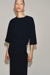 Vestido midi de cuello redondo y manga corta, confeccionado en tejido fluido color azul marino. Presenta un diseño con detalle de drapeado y volado lateral en la parte delantera, con cierre en la espalda mediante abertura y botón.