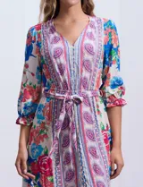 Vestido midi con estampado floral en tonos azules, rojos y blancos. Tiene escote en V, mangas 3/4 con puños fruncidos, cintura elastizada con lazo de ajuste y falda con volantes.