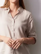 Camisa color beige de tencel, marca Hester & Orchand, con cuello con solapa, escote en V, abertura central con botones y bolsillos delanteros. Las mangas son largas y regulables con presilla y botón.