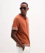 Camisa de punto jacquard color naranja, con cuello solapa, manga corta y cierre frontal con botones. Presenta detalles de rayas horizontales en contraste en el ruedo y en los puños de las mangas.